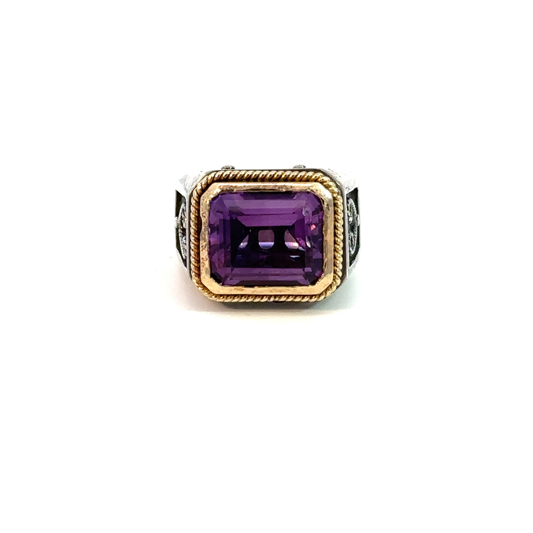 A Konstantino Late Century ring featuring an emerald-cut, 5-carat amethyst set in the center. The amethyst is framed by 18kt yellow gold accents, and the sterling silver band is adorned with intricate black detailing. This ring is displayed upright against a white background.