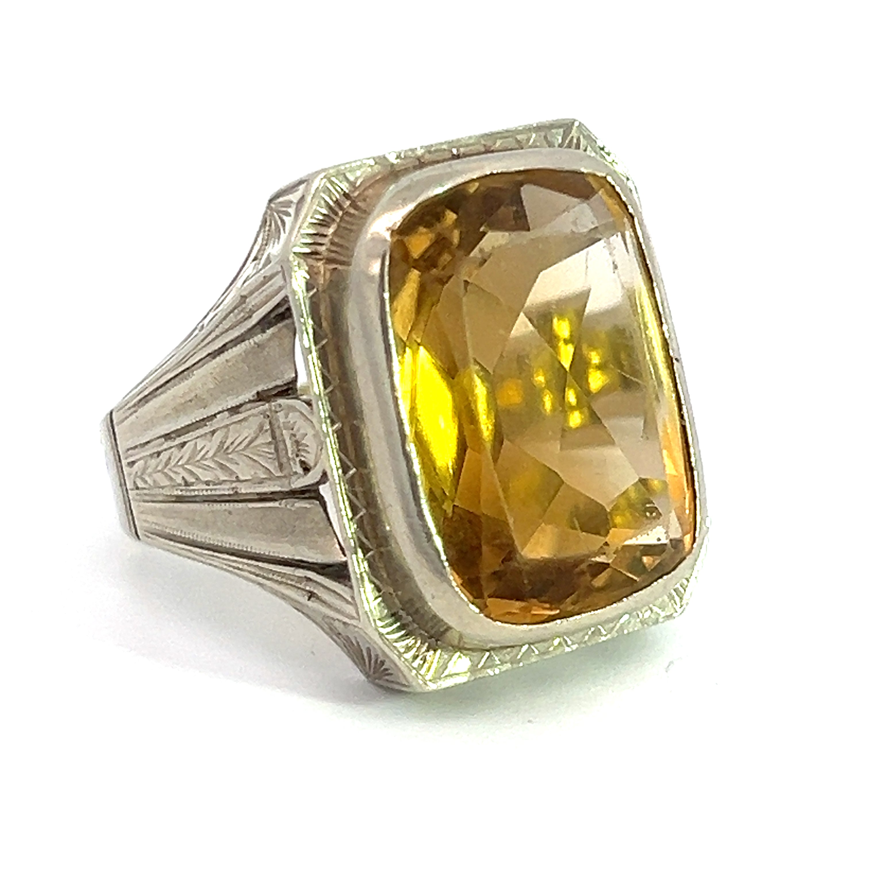 A close-up of an Art Deco ring showcasing a 15ct citrine gemstone set in a finely engraved 14kt white gold band. The slightly cushion-shaped, rectangular-cut gem has beveled edges and reflects light, displaying various shades of yellow and gold.