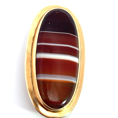 A close-up image of an oval banded agate stone with horizontal stripes in shades of brown, white, and red. Set in a gold-toned frame, the Victorian 10ktyg Banded Agate Pin exudes a polished and elegant appearance that evokes the charm of antique Victorian jewelry. The background is plain white.
