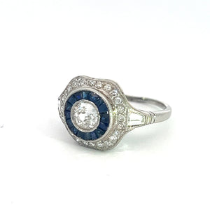 The Art Deco Platinum .65ct Old Mine Cut Diamond + 1cttw Sapphires + 1.50cttw Diamonds Ring boasts an intricate design with a prominent large central old mine cut diamond, encircled by smaller blue sapphires and an outer band of tiny diamonds. This vintage-style ring is elegantly displayed against a plain white background.