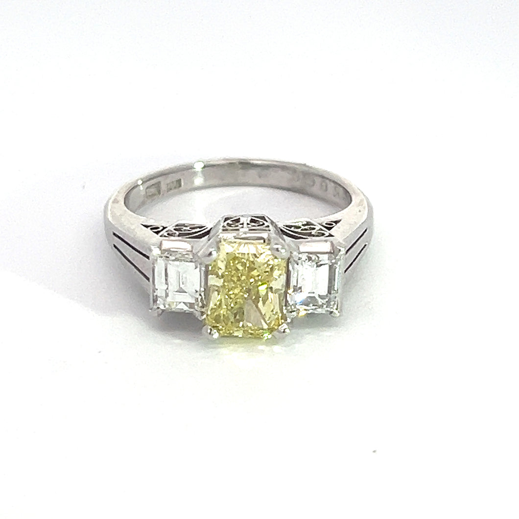 The Modern Platinum 1.29ct Fancy Yellow Radiant Cut Diamond + .66cttw Emerald Cut Diamond Ring showcases a stunning 1.29ct radiant cut fancy yellow diamond at its center, complemented by two rectangular clear emerald cut diamonds on either side. The ring band features sleek and elegant cuts, enhancing its overall sophistication.