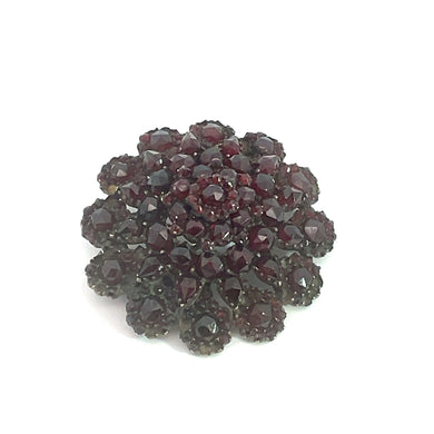 The Victorian Vermeil Garnet Wreath Style Pin is a circular, multi-layered brooch showcasing dark red, faceted garnets arranged in a flower-like pattern against a white background. This piece of Victorian era jewelry exudes vintage charm with its intricate design and several layers of arranged gemstones, creating a captivating three-dimensional effect.