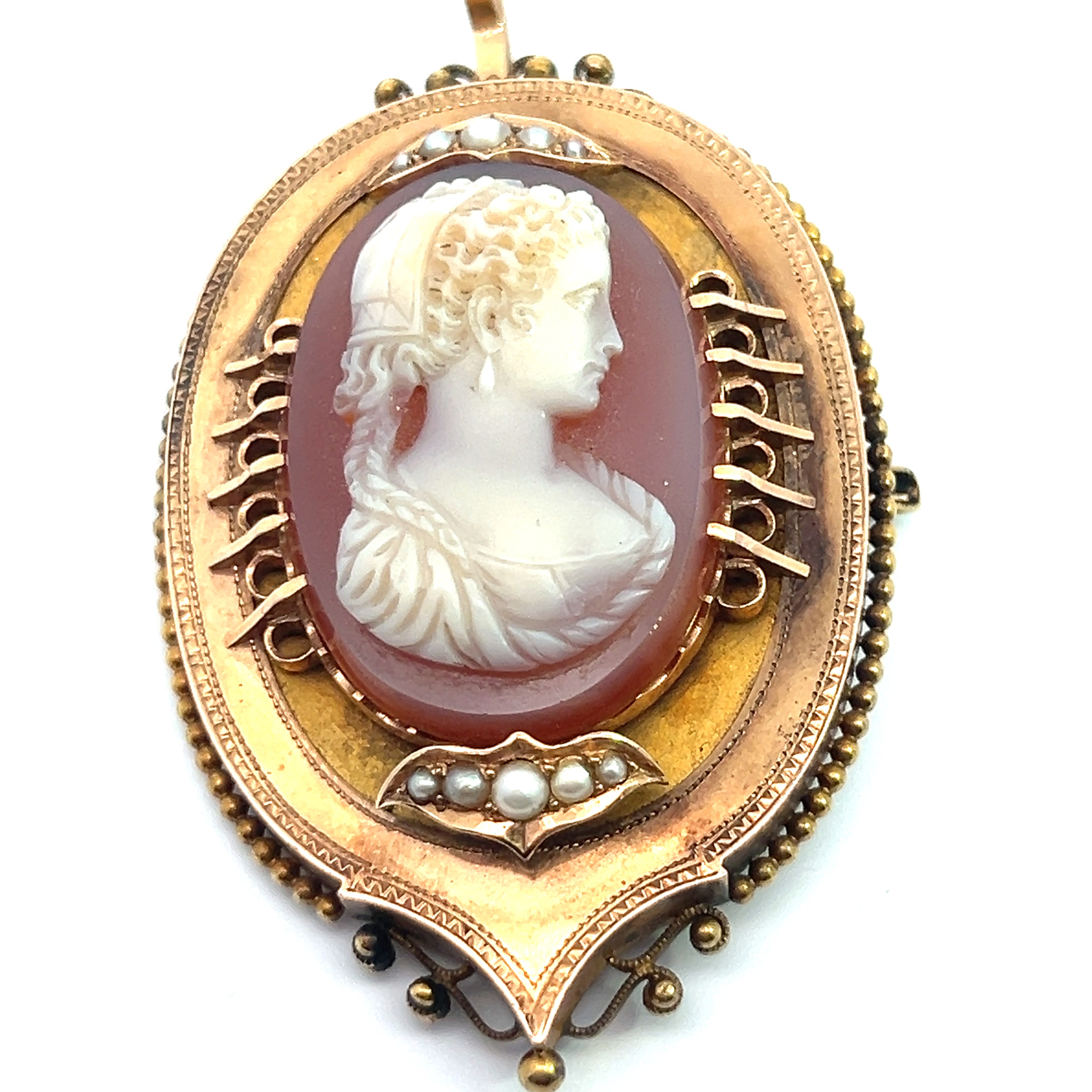The Victorian 14ktyg Carved Agate Portrait of a Lady Cameo is an antique brooch showcasing a carved agate portrait cameo of a woman in relief against a reddish-pink background. The frame features intricate 14kt yellow gold filigree and is adorned with small pearls, creating a timeless and elegant design. The brooch is equipped with a pin and clasp at the back.
