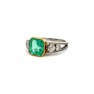 The Mid Century Platinum & 18kt Yellow Gold 4.03 Emerald Cut Emerald + .70cttw Diamond Ring showcases an elegant design with a large, 4.03ct emerald-cut green gemstone set in a gold bezel. The sophisticated platinum band is adorned with three round diamonds on each side of the central stone, exuding Mid Century flair, and is photographed against a plain white background.