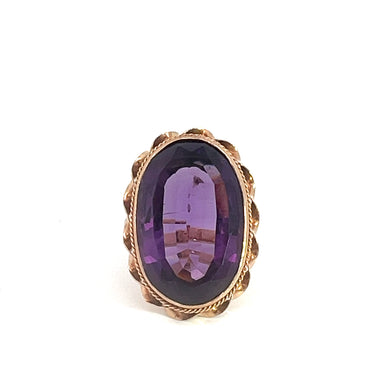 A close-up photograph of the Victorian 14kt Yellow Gold 24ct Oval Amethyst Fancy Framed Ring with a stunning oval-shaped, faceted purple gemstone set in the center. The gemstone, a magnificent 24ct oval amethyst, is framed by an intricate gold setting that features a braided border design. The ring exudes vintage charm and is positioned vertically against a white background.