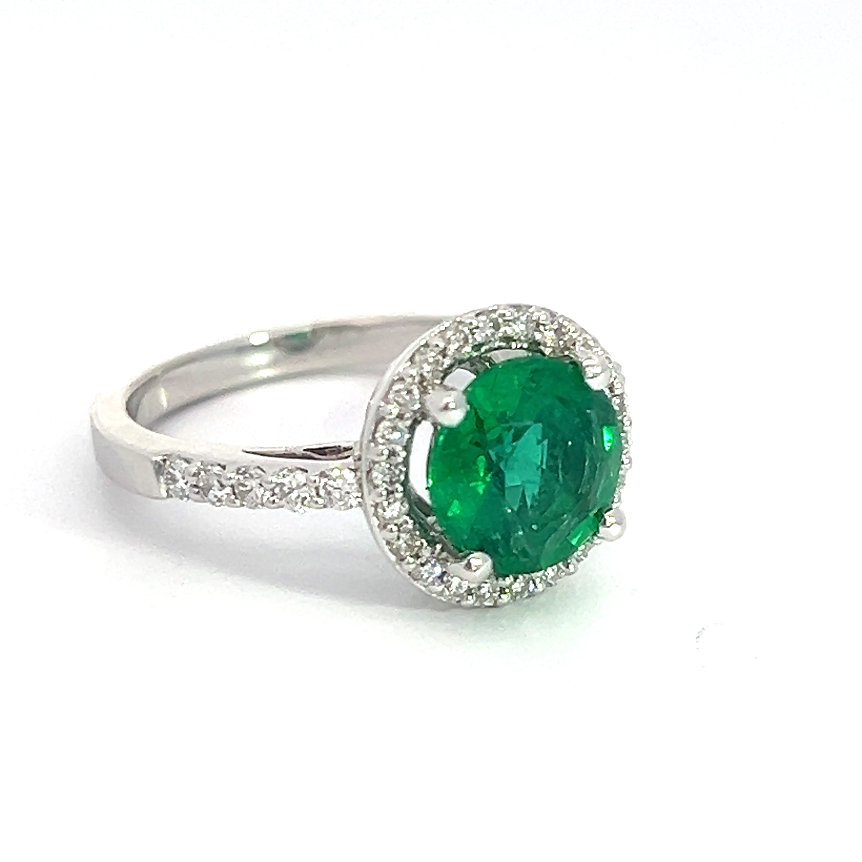 The Modern 18kt White Gold 1.80ct Emerald + .50cttw Diamond Ring showcases a stunning silver band adorned with small diamonds, creating a sparkling, elegant appearance against a plain white background. This exquisite piece features a large, 1.80ct round emerald at its center, surrounded by a halo of .50cttw small white diamonds and is crafted from luxurious 18kt white gold.