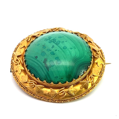 The Victorian 18ktyg Fancy Frame Malachite Brooch showcases a large, polished malachite stone with natural patterning, set in a detailed gold frame adorned with leaf motifs. The green malachite stone is prominently centered, showcasing its unique swirls and speckles. The brooch is displayed on a plain white background.