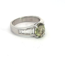 Load image into Gallery viewer, A Late Century Platinum ring featuring a central 3.03ct oval yellow-green Alexandrite gemstone. With a sleek design, the band showcases 1.28cttw small rectangular (baguette cut) white diamonds embedded alongside the central stone, creating an elegant contrast against the plain white background.