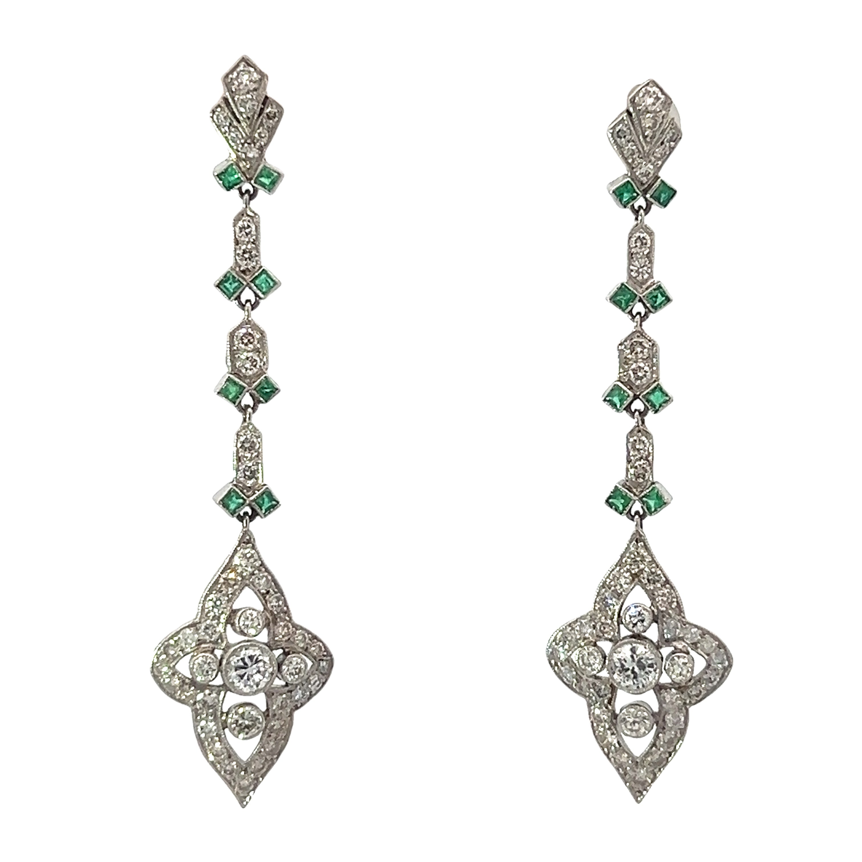 A pair of Late Art Deco 4.50cttw Round Brilliant Cut Diamonds + .80cttw Emeralds Dangle Earrings, featuring a floral design with sparkling round brilliant cut diamonds and small green emerald accents. Each platinum earring showcases an intricate pattern with a central flower motif and a series of geometric shapes connecting the elements.