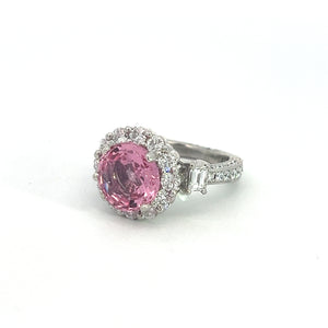 The Modern Platinum 3.30 No Heat Padparadscha Sapphire Ring features an exquisite, rare gemstone as its centerpiece: a large, round pink no-heat Padparadscha sapphire surrounded by smaller clear diamonds in a halo setting. The band is adorned with additional clear diamonds and rectangular side stones.