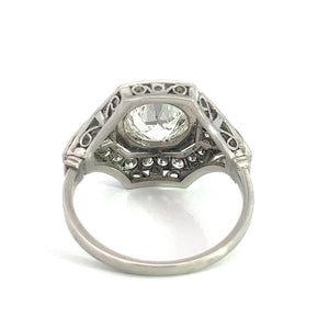 The Art Deco Platinum 2ct Old European Cut Diamond + .84cttw Diamond Ring features an ornate, intricate filigree design on the sides, showcasing a large, round-cut gemstone set atop it. This ring's detailed craftsmanship includes small decorative accents beneath the main stone. The vintage diamond ring is displayed against a white background.