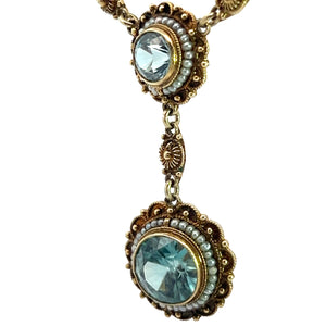Introducing the Victorian 14kt Yellow Gold 5.50cttw Blue Zircon + Seed Pearls Pendant: a vintage-style necklace showcasing two stunning circular blue zircons, encircled by intricate gold detailing and accented with delicate seed pearls. The larger zircon elegantly dangles beneath the smaller one, both linked by a gold connector embellished with additional filigree designs.