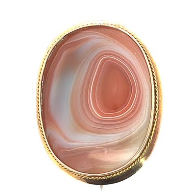 The Victorian 14ktyg Large Banded Agate Pin is a vintage oval brooch featuring a polished, multi-layered pink agate stone, encased in a 14kt yellow gold setting adorned with an elegant twisted rope design along the edges. This exquisite jewel from the Victorian era showcases varying shades of pink, white, and subtle traces of light brown.
