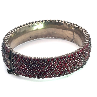 The Victorian Vermeil Garnet Bangle Bracelet is adorned with small, round red stones that cover the entire outer surface, exuding vintage elegance. Its smooth, reflective interior complements the textured inner edge and a secure clasp allows for easy opening and closing, embodying Victorian sophistication.