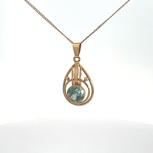 An Art Nouveau 14kt yellow gold necklace with a delicate chain featuring a teardrop-shaped pendant. The pendant has an open geometric design and holds a single, round blue zircon gemstone at its center. The background is white.