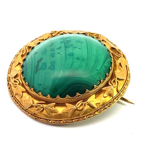 Photograph of the Victorian 18ktyg Fancy Frame Malachite Brooch showcasing a large, round, green malachite gemstone set in an ornate gold-toned metal frame. The frame is adorned with intricate leaf patterns and delicate scrollwork. The brooch is displayed on a plain white background.