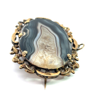 An intricate Victorian 10ktyg banded agate and quartz crystal pendant/pin featuring an oval-shaped, banded agate stone set in a beautifully detailed gold-toned frame. Reflecting the elegance of Victorian jewelry, this piece comes with a pin clasp on the back. The agate stone showcases natural banded patterns in enchanting shades of gray, white, and tan.