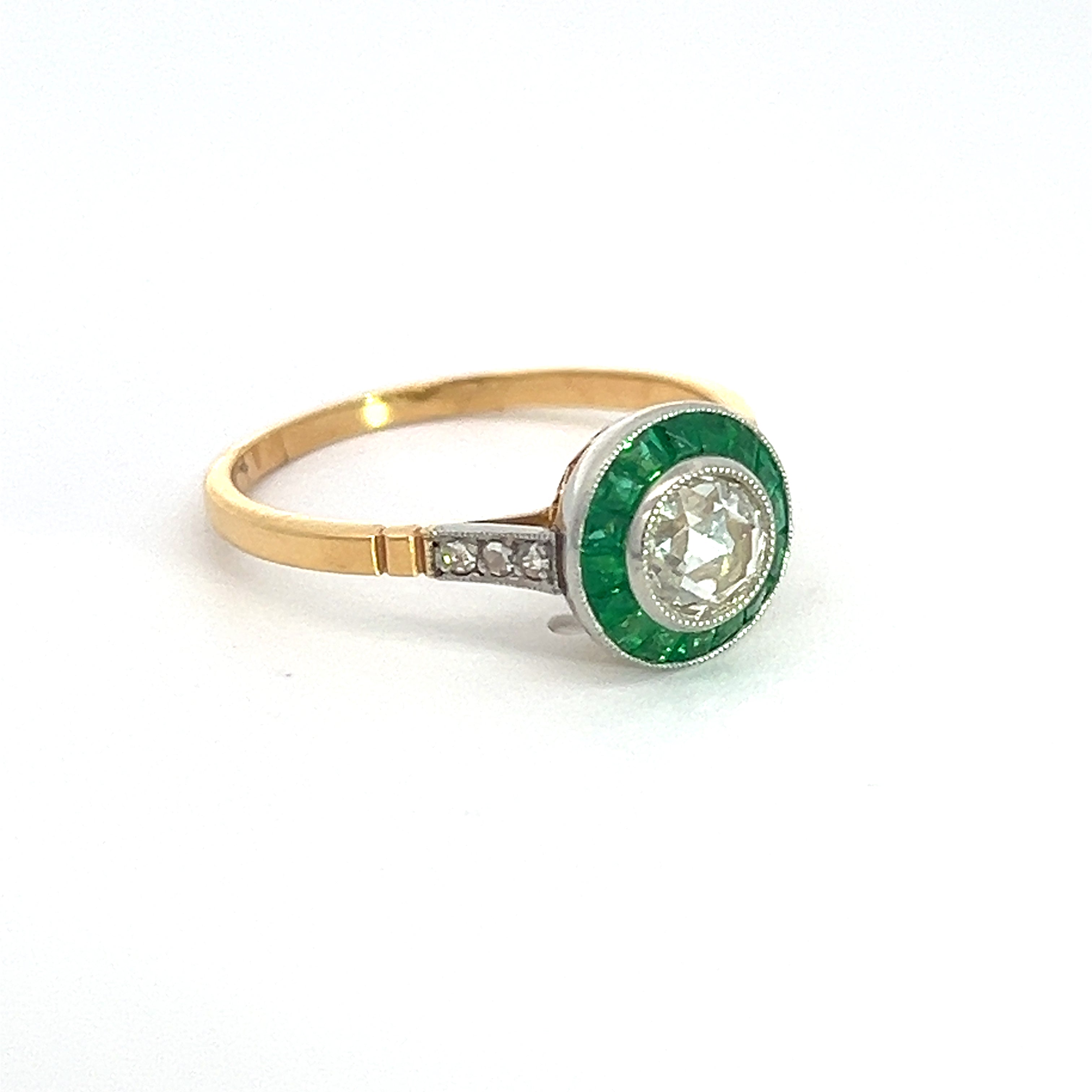 Presenting the Edwardian Platinum/14KYG Ring, a stunning piece featuring a .70ct rose-cut diamond at the center, gracefully encircled by a 1cttw halo of emeralds. The band combines tones of gold and platinum and is delicately embellished with an additional .15cttw of diamonds on either side of the central emerald halo.