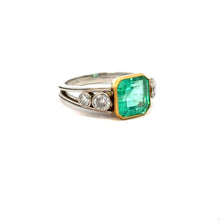 Load image into Gallery viewer, The Mid Century Platinum &amp; 18kt Yellow Gold 4.03 Emerald Cut Emerald + .70cttw Diamond Ring features a central, vibrant green emerald weighing 4.03ct flanked by two round diamonds on each side. The emerald is set in a gold bezel setting, contrasting against the silver band. This ring exudes modern elegance with mid-century sophistication.