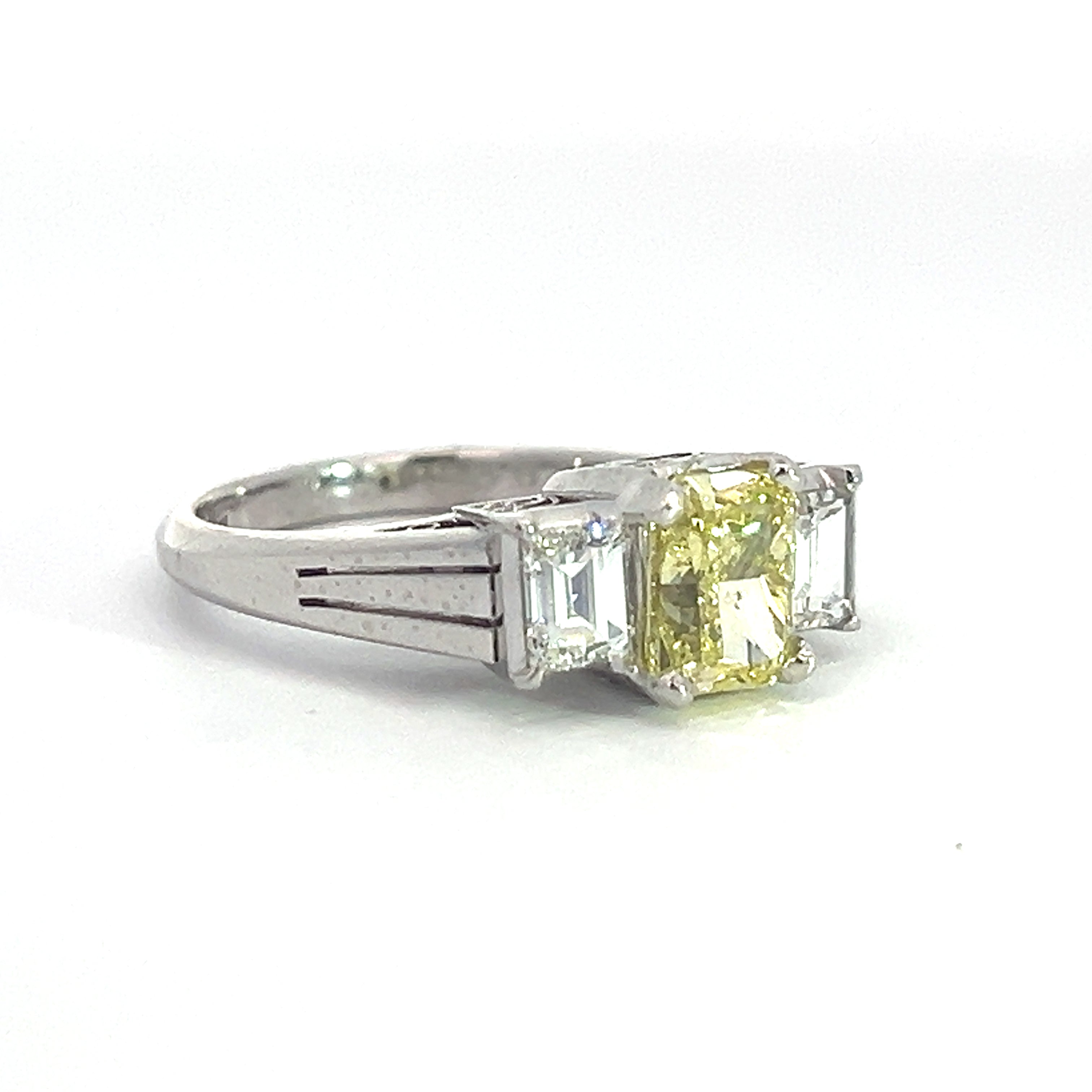 The Modern Platinum 1.29ct Fancy Yellow Radiant Cut Diamond + .66cttw Emerald Cut Diamond Ring showcases a stunning 1.29ct radiant cut fancy yellow diamond at its center, complemented by two rectangular clear emerald cut diamonds on either side. The ring band features sleek and elegant cuts, enhancing its overall sophistication.