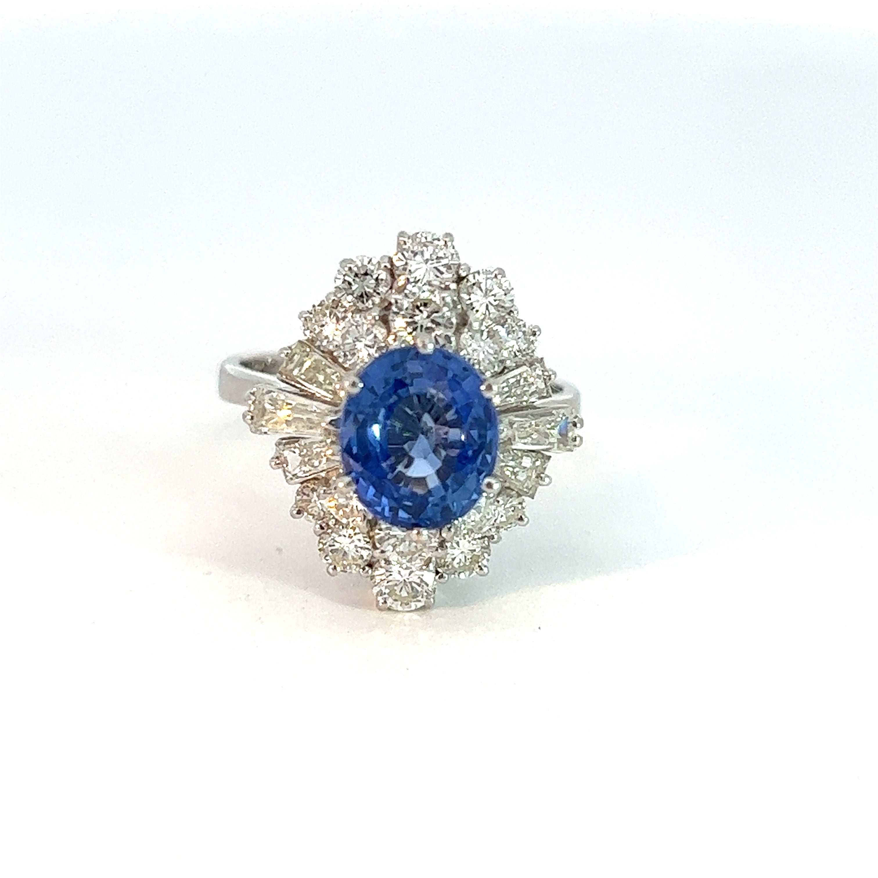 Introducing the Mid Century 18kt White Gold 2.56 Sapphire + 2.26ct Diamond Ring: a breathtaking piece featuring a large, oval-shaped blue sapphire gemstone encircled by a halo of dazzling, clear diamonds. The intricate design and 18k white gold band enhance its brilliance, making it an exquisite example of luxury jewelry.
