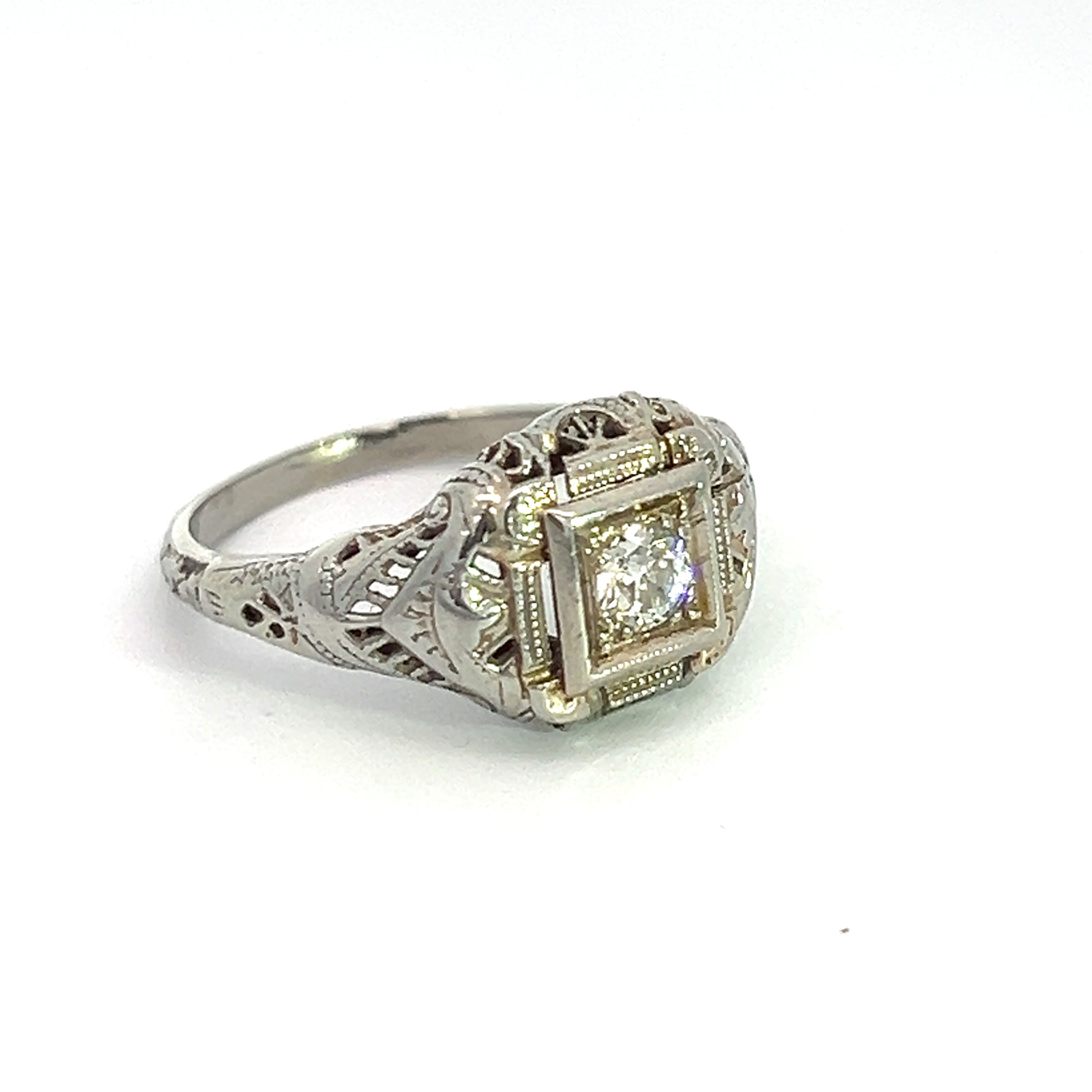 A captivating piece, the Art Deco 18ktwg .20ct Old Mine Cut Diamond Ring boasts an exquisite filigree design in 18kt White Gold. The centerpiece is a single round Old Mine Cut Diamond securely set within a square bezel on the front. Displayed gracefully against a plain white background, this ring exudes timeless elegance.