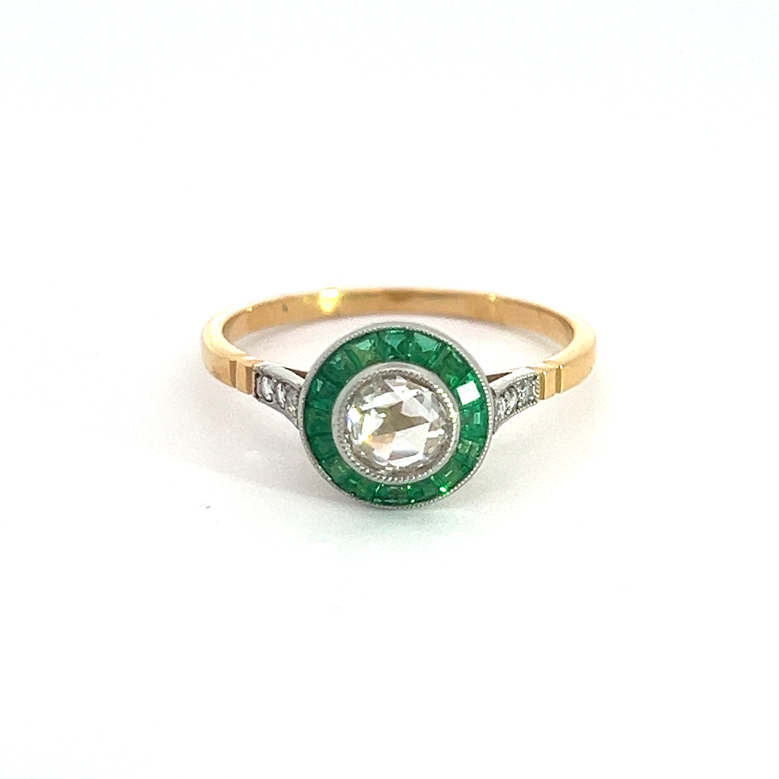 Presenting the Edwardian Platinum/14KYG Ring, a stunning piece featuring a .70ct rose-cut diamond at the center, gracefully encircled by a 1cttw halo of emeralds. The band combines tones of gold and platinum and is delicately embellished with an additional .15cttw of diamonds on either side of the central emerald halo.