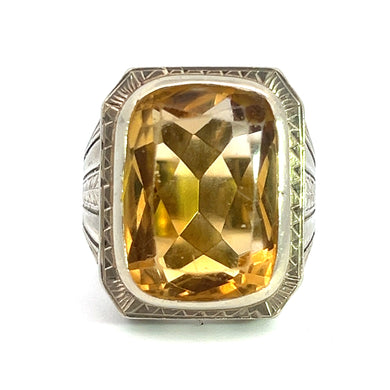 A close-up of an Art Deco ring showcasing a 15ct citrine gemstone set in a finely engraved 14kt white gold band. The slightly cushion-shaped, rectangular-cut gem has beveled edges and reflects light, displaying various shades of yellow and gold.