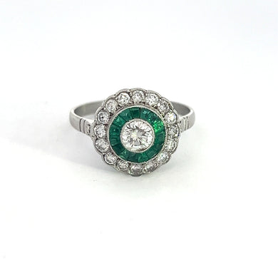 A silver ring featuring a large central diamond surrounded by a circle of green emerald gemstones and an outer circle of smaller diamonds, embodying vintage elegance. The Late Art Deco .35ct Round Brilliant Cut Diamond + .85cttw Diamonds + .60ct Emerald Halo Ring is set against a plain white background, showcasing its timeless allure with Late Art Deco charm.