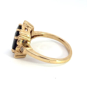 A Mid Century 14kt yellow gold ring, named the "Mid Century 14ktyg 4.80ct Garnet + .06cttw diamond ring," features an emerald-cut dark garnet gemstone set within a prong setting and adorned with smaller accent diamonds on the shoulders. The ring, showcased from a side angle and resting on a white surface, exudes vintage charm.