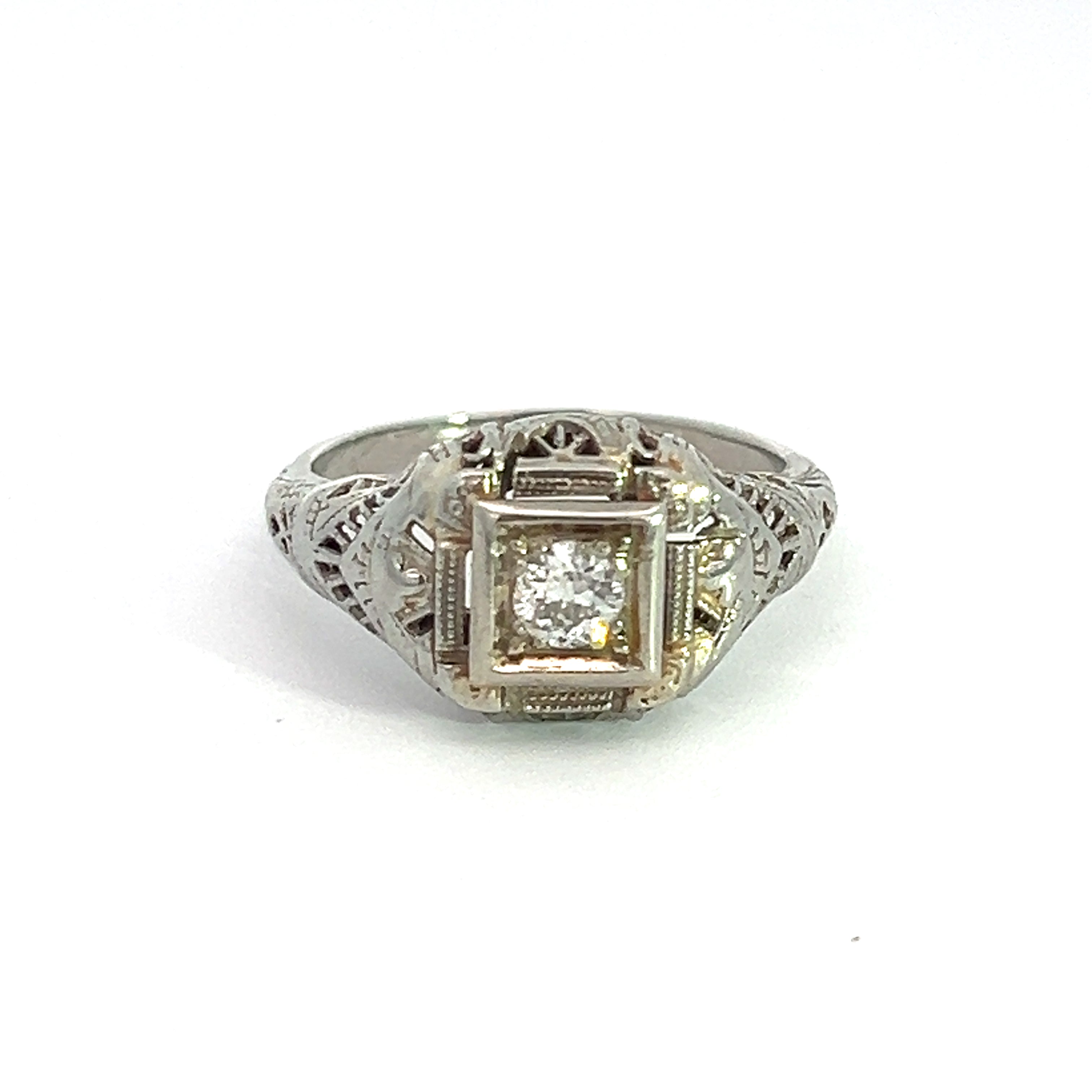 A captivating piece, the Art Deco 18ktwg .20ct Old Mine Cut Diamond Ring boasts an exquisite filigree design in 18kt White Gold. The centerpiece is a single round Old Mine Cut Diamond securely set within a square bezel on the front. Displayed gracefully against a plain white background, this ring exudes timeless elegance.