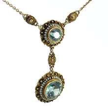 Load image into Gallery viewer, The Victorian 14kt Yellow Gold 5.50cttw Blue Zircon + Seed Pearls Pendant showcases a vintage-style design, featuring two round blue gemstones set in intricate gold-toned settings with small pearls around the edges. The gems are linked together vertically, and the chain is adorned with detailed gold-toned ornamental designs.