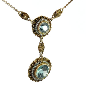 The Victorian 14kt Yellow Gold 5.50cttw Blue Zircon + Seed Pearls Pendant showcases a vintage-style design, featuring two round blue gemstones set in intricate gold-toned settings with small pearls around the edges. The gems are linked together vertically, and the chain is adorned with detailed gold-toned ornamental designs.