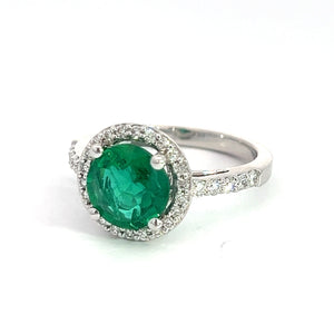 Introducing the Modern 18kt White Gold 1.80ct Emerald + .50cttw Diamond Ring, showcasing a stunning 1.80ct emerald at its center, encircled by a halo of small white diamonds totaling .50cttw. The band is elegantly adorned with additional small white diamonds on the sides, further enhancing its sophisticated and luxurious look.