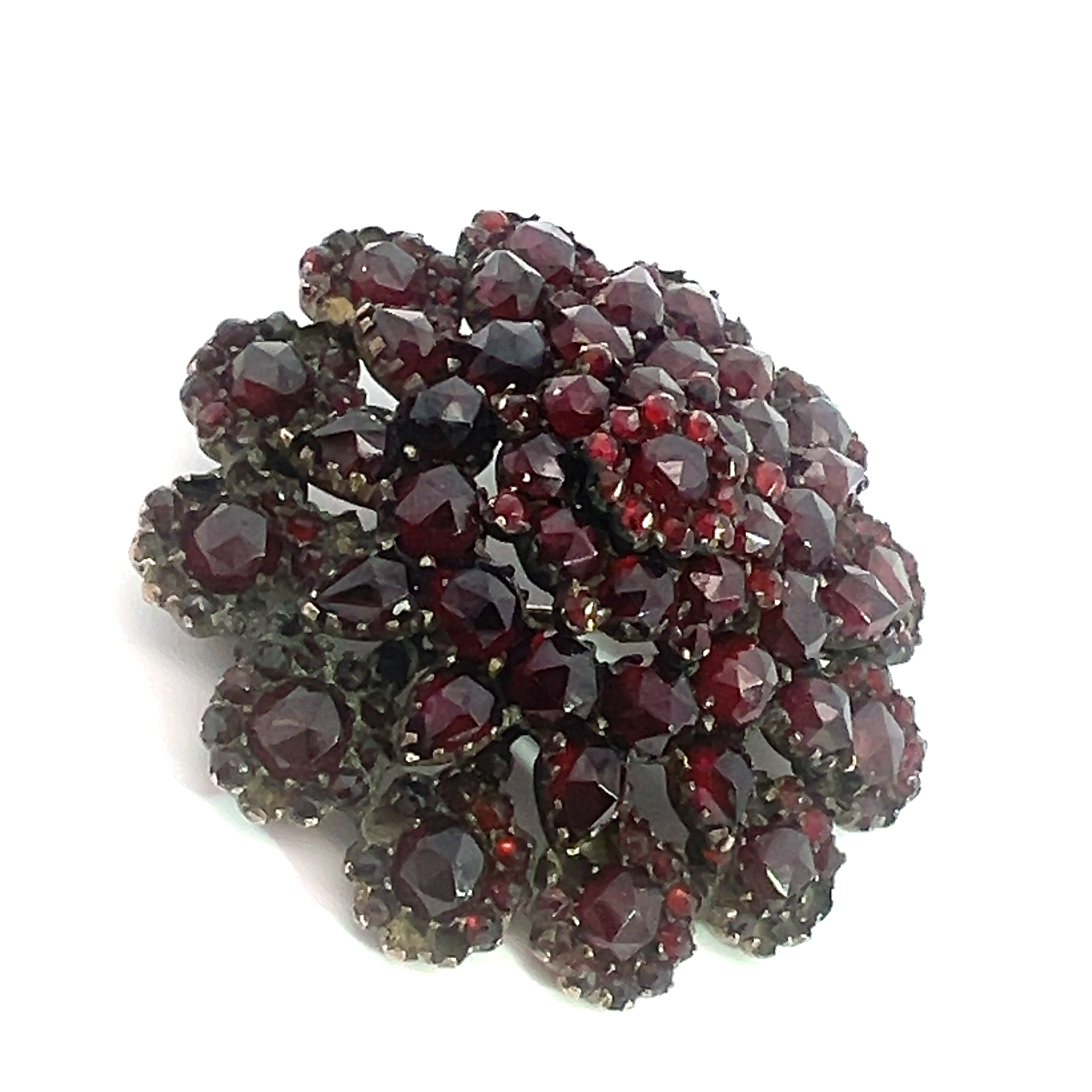 The Victorian Vermeil Garnet Wreath Style Pin is a circular, multi-layered brooch showcasing dark red, faceted garnets arranged in a flower-like pattern against a white background. This piece of Victorian era jewelry exudes vintage charm with its intricate design and several layers of arranged gemstones, creating a captivating three-dimensional effect.