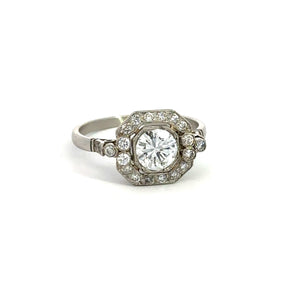 The Art Deco Platinum .80ct Round Brilliant Cut + 1.08cttw Diamond Ring showcases an elegant central round brilliant cut diamond encircled by smaller diamonds arranged in a geometric pattern. The band is simple, enhancing its sophisticated and classic look. The ring is displayed against a plain white background, emphasizing its beauty.