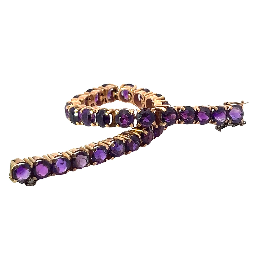 A Late Century 14kt Yellow Gold 14cttw Amethyst Tennis Bracelet, adorned with alternating round purple gemstones, is partially coiled to showcase the sparkling facets against a plain white background.