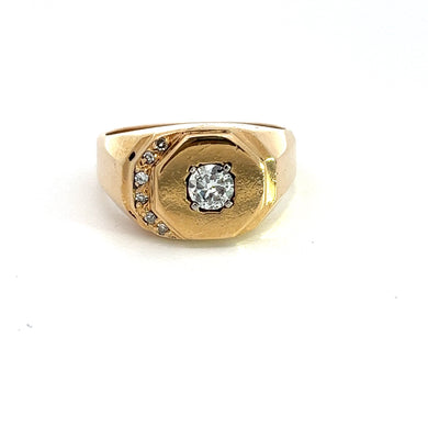 The Art Deco 14kt Yellow Gold .40ct Round Brilliant + .10cttw Diamond Ring features a central round brilliant diamond set in an octagonal bezel. The band elegantly showcases smaller diamonds arranged diagonally on one side, creating a sophisticated and elegant design against a white background.