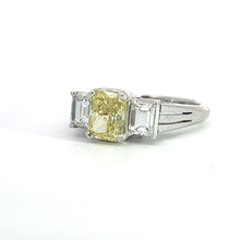 Load image into Gallery viewer, The Modern Platinum 1.29ct Fancy Yellow Radiant Cut Diamond + .66cttw Emerald Cut Diamond Ring features a radiant cut fancy yellow diamond of 1.29 carats, flanked by two smaller clear emerald cut diamonds totaling .66 carats, set against a white background. The ring showcases a sleek and elegant design, with all stones securely held by prongs.