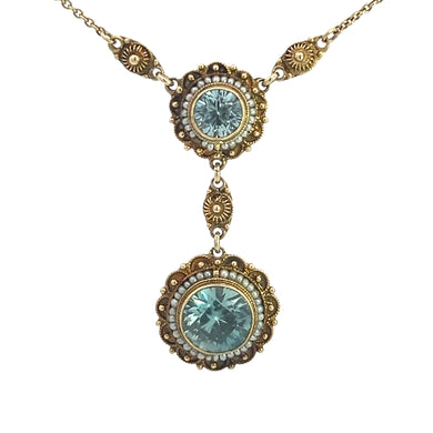 The Victorian 14kt Yellow Gold necklace showcases two ornate pendants with intricate designs. Each pendant features a large, round blue zircon gemstone at the center, surrounded by smaller seed pearls. The upper pendant is elegantly connected to the lower pendant by a small decorative link.
